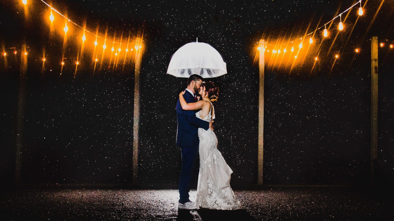 bride groom kissing under clear umbrella rain with cafe lights