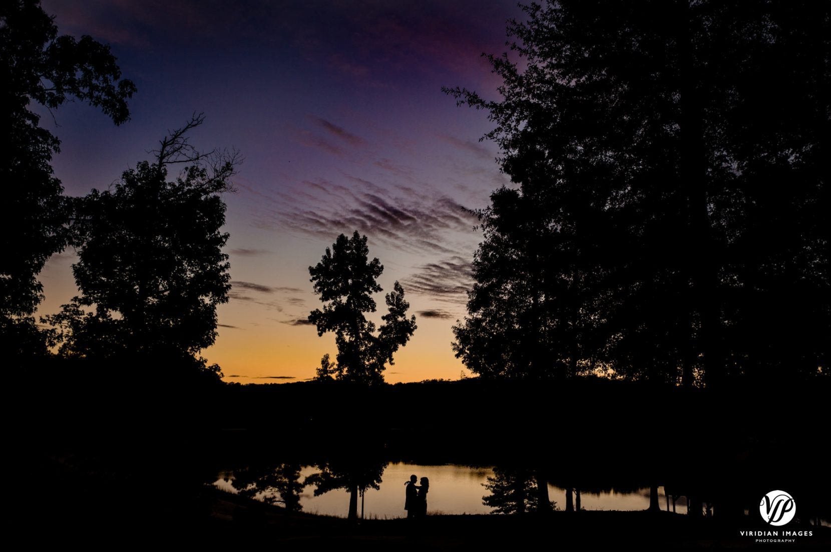 wide angle shot of couple silhouette against mirror reflected lake at sunset