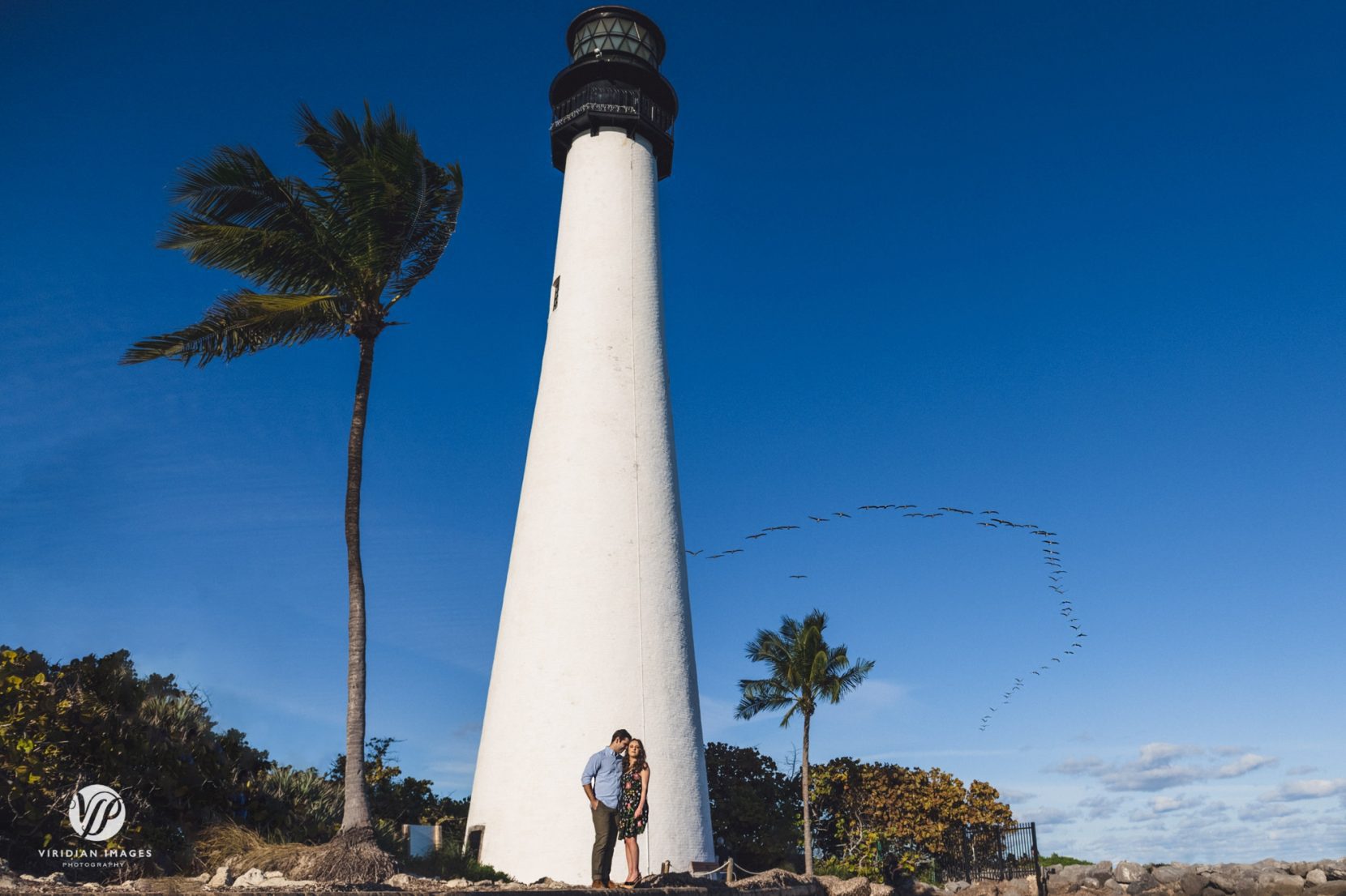 couple bottom of white lighthouse with flock of birds flying above in the blue sky