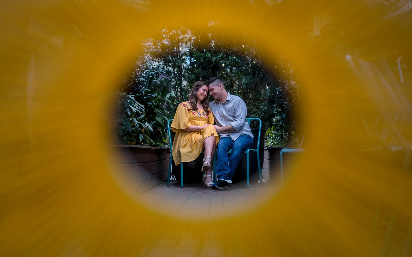 couple sitting on teal chairs foreheads touching and smiling. shot through bright yellow weaved chair
