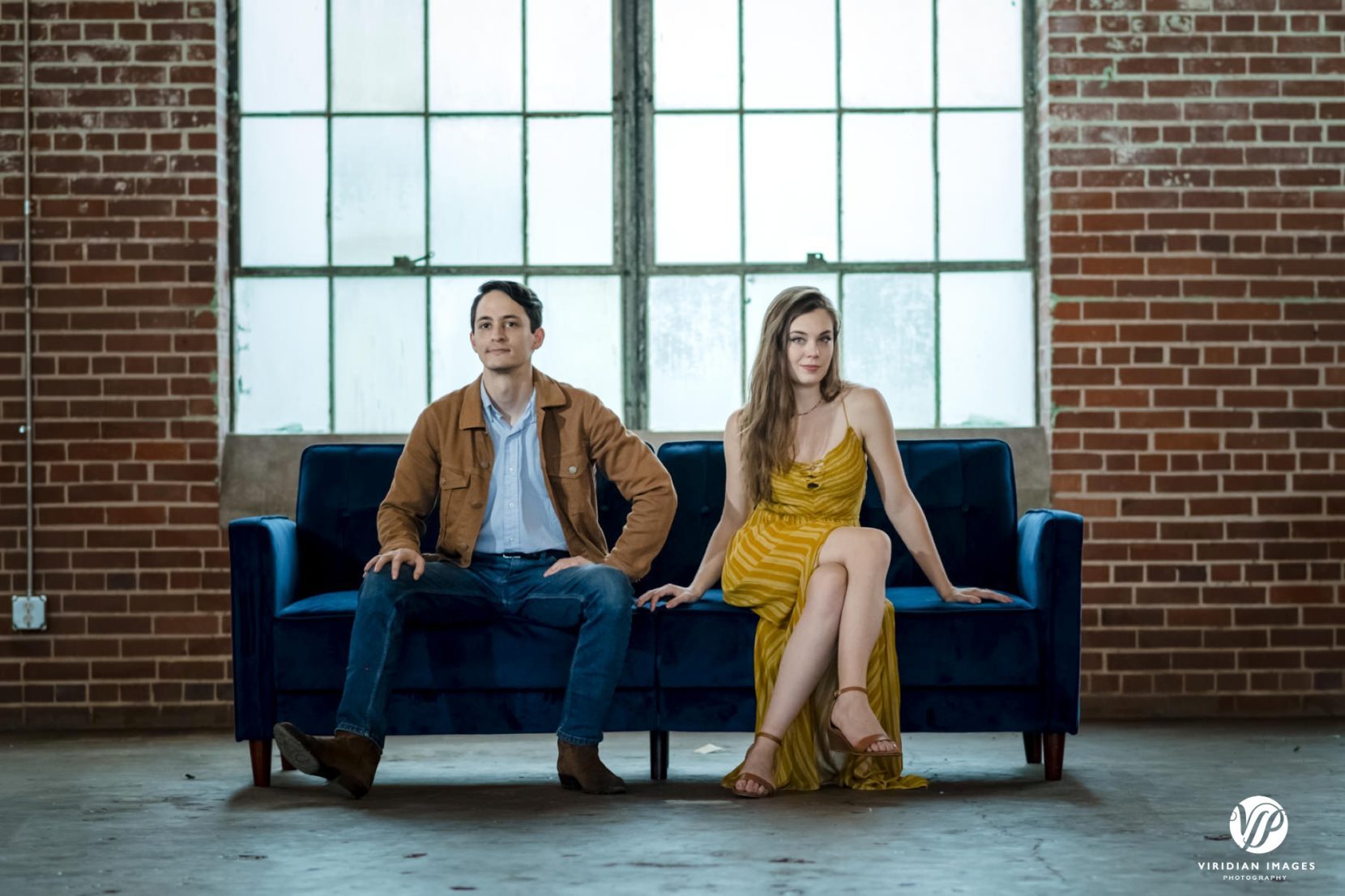 Editorial style couple photo on blue sofa in front of industrial windows during engagement session at Lindale Mill