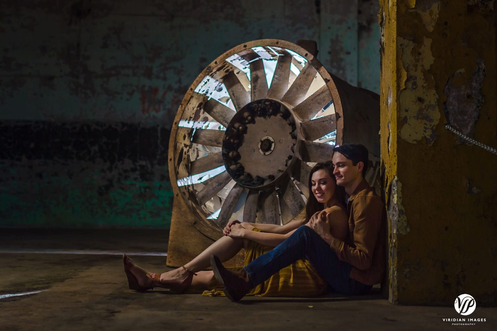 Couple sitting in front of back-lit turbine on factory floor at Lindale Mill during engagement session