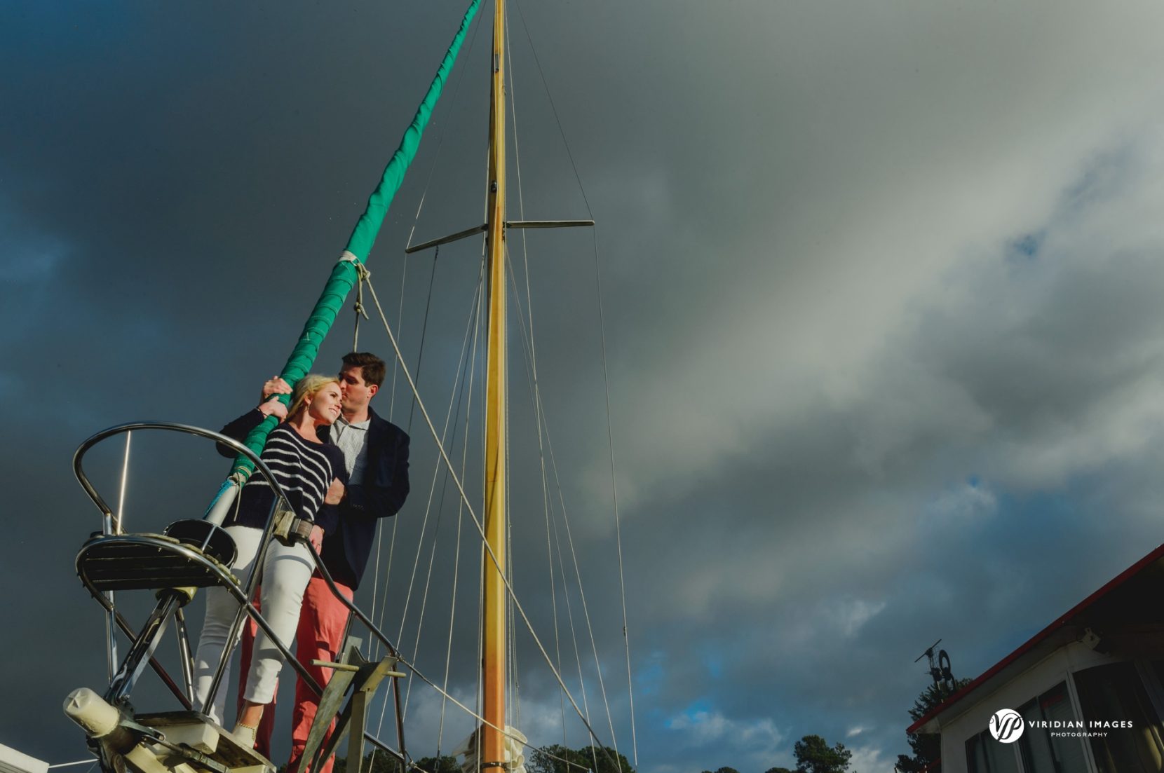 Couple standing on bow of sailboat with dark stormy clouds behind during winter photoshoot