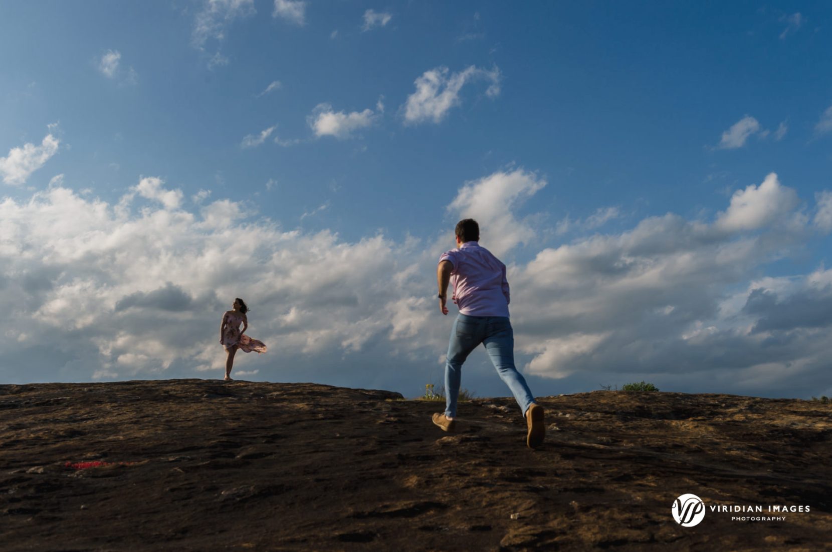 Wide shot of groom-to-be running towards bride against blue skies and clouds during spring engagement shoot on top of Arabia Mountain