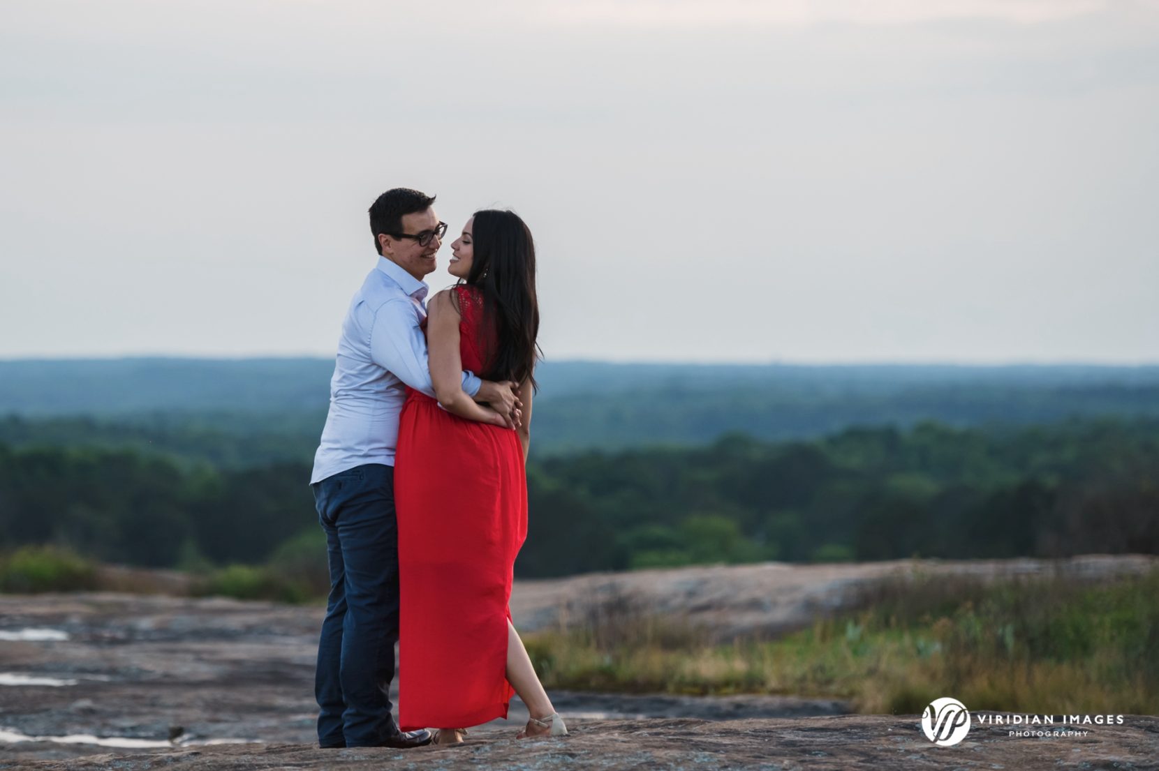 man holds woman in red dress atop of mountain with tree filled horizon