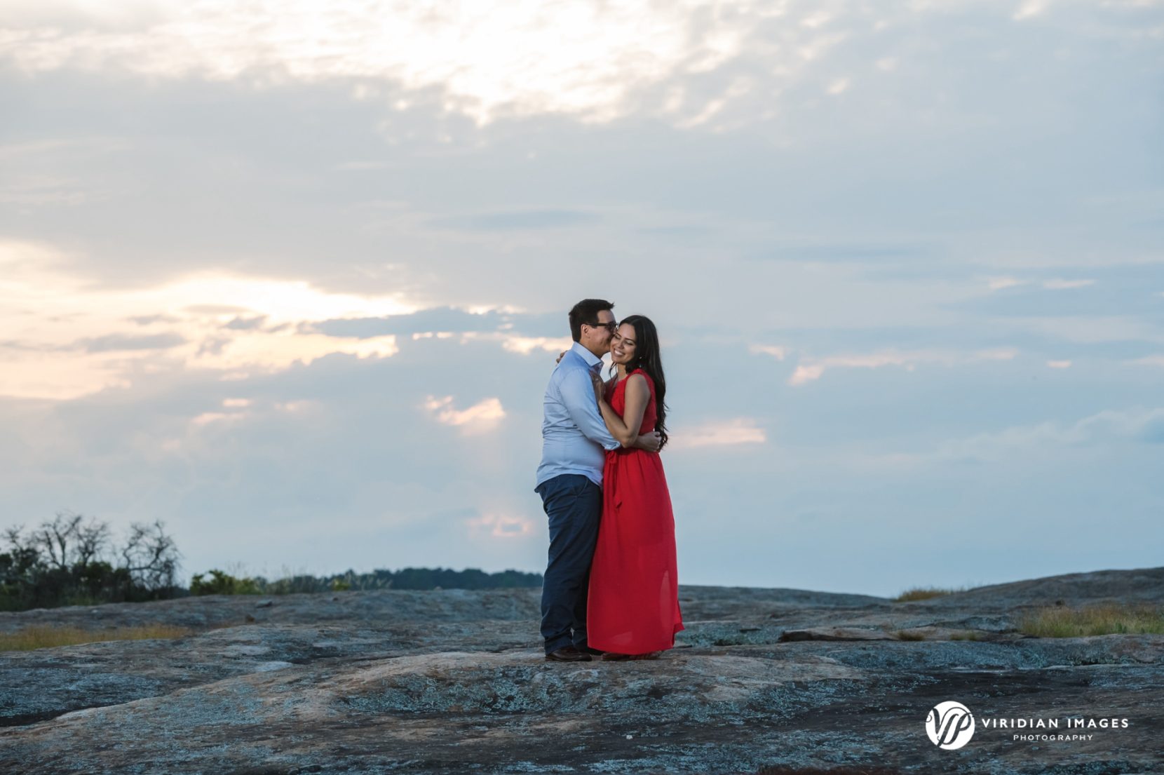 man kisses woman in red dress against sunset filled sky and clouds during engagement session
