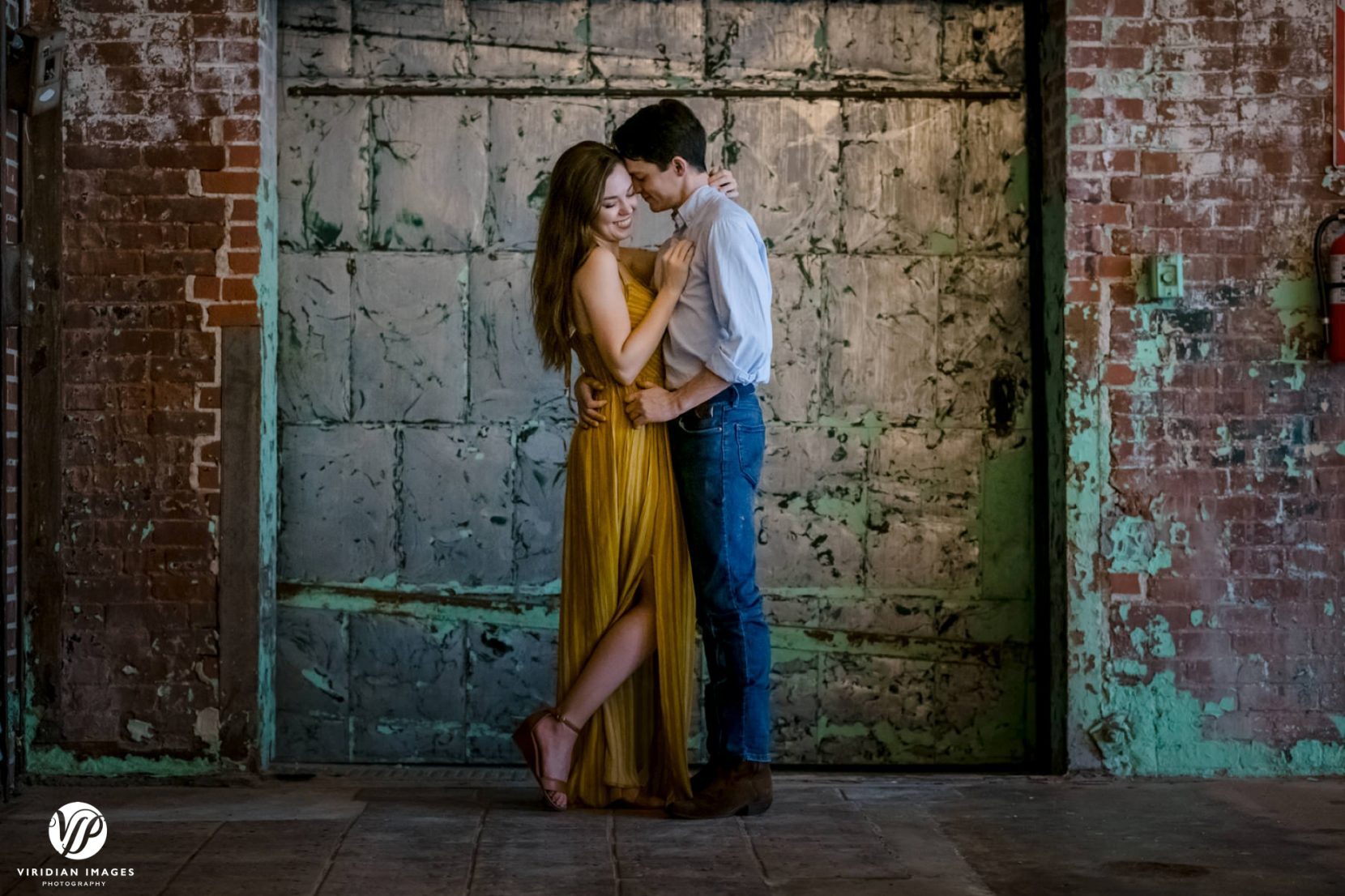 Couple embracing in front of rusted factory doors during engagement session at Lindale Mill