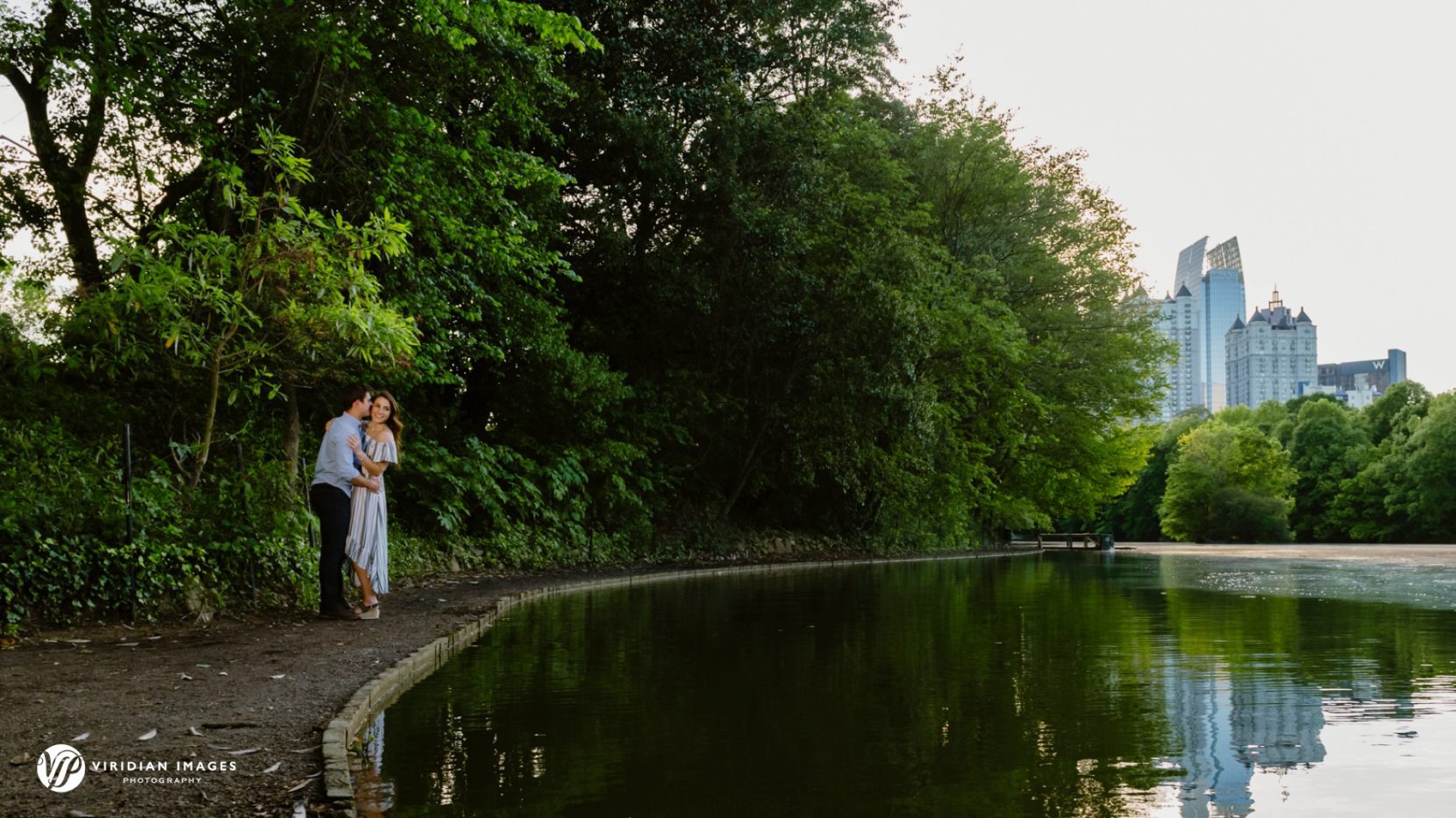 Wide shot of couple walking along lake with city skyline at Piedmont Park engagement session 