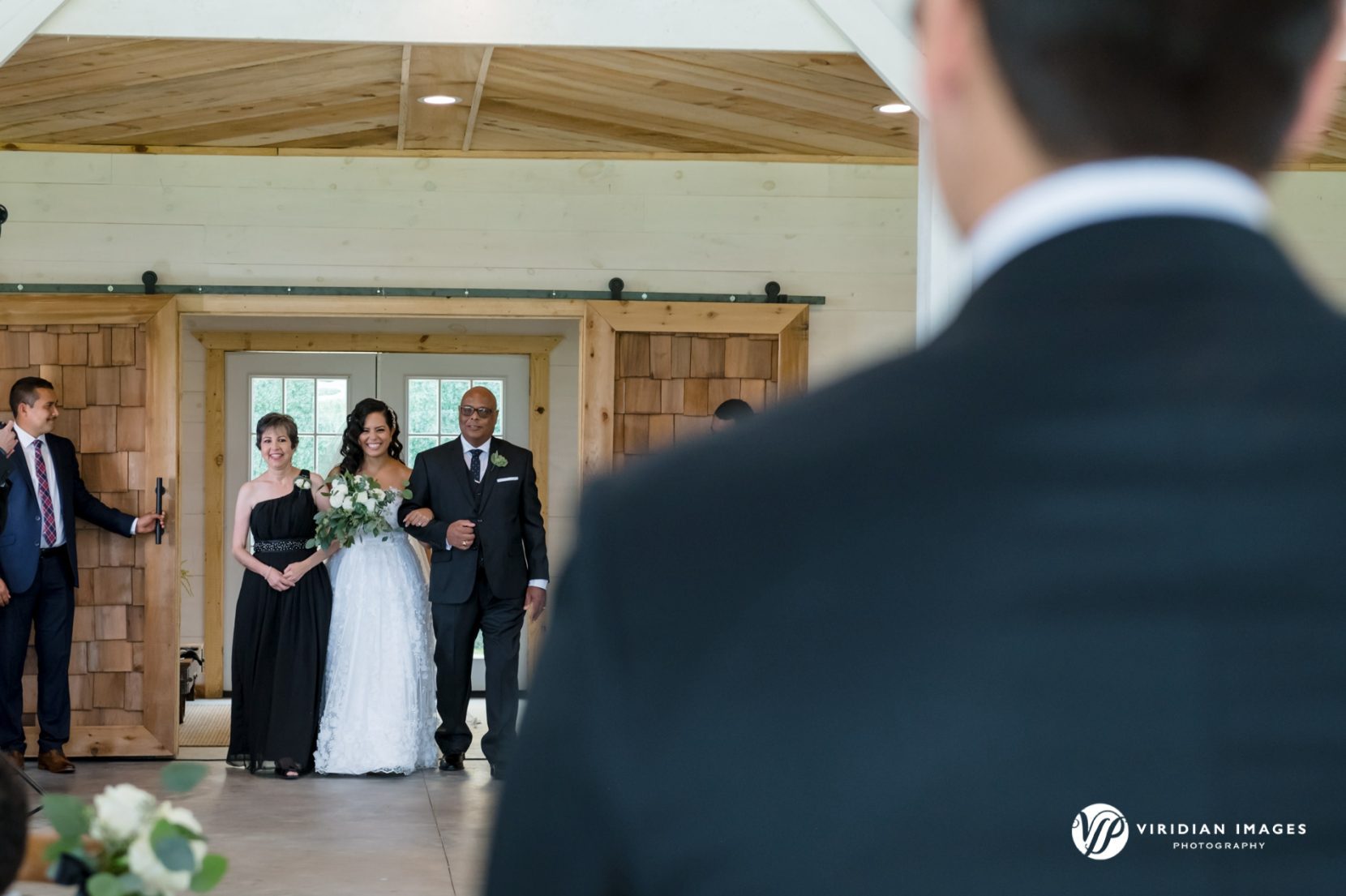 Bride with mom and dad enter through double doors during wedding processional groom at foreground 