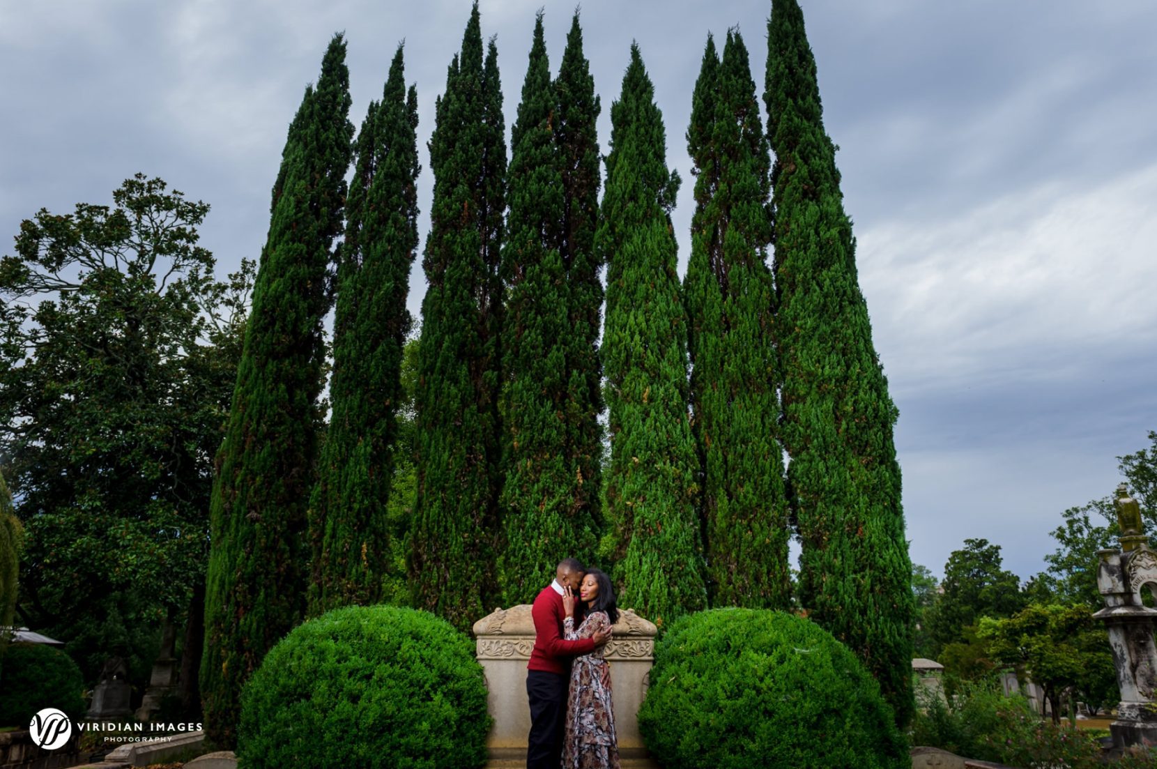 Wide photo of engaged couple in front of cypress tress embracing at Oakland Cemetery