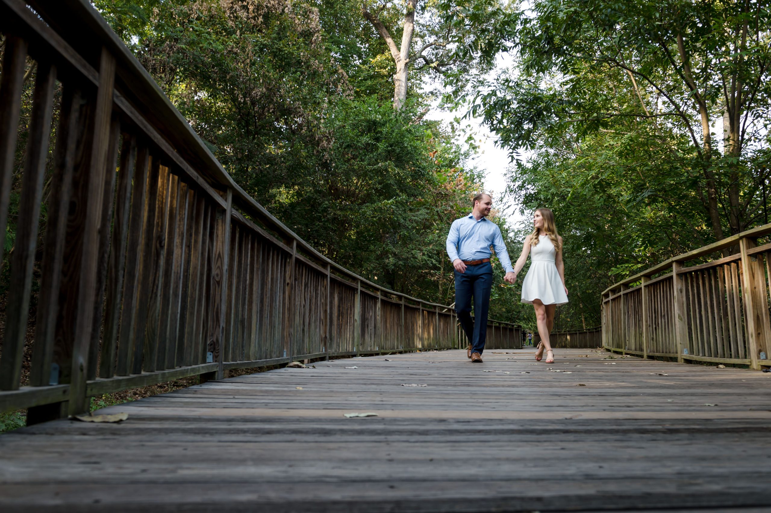 Engaged couple walking through wood pathway looking at each other