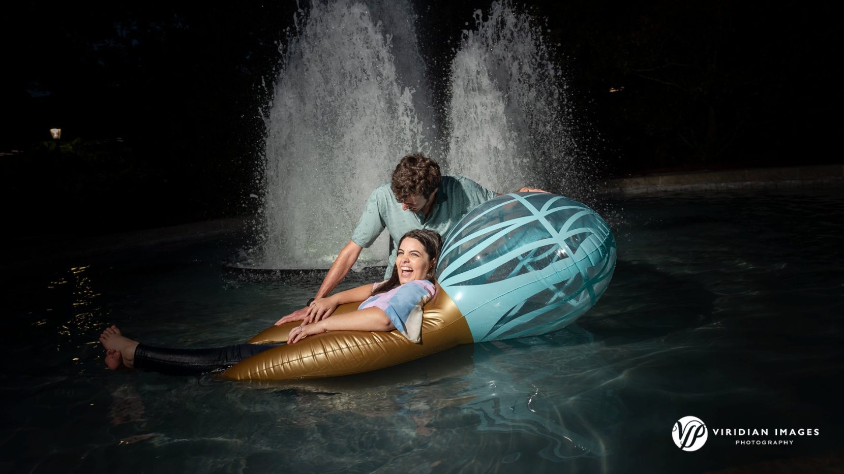 Engaged couple laughing while floating on inflatable ring in fountain at UGA North Campus.