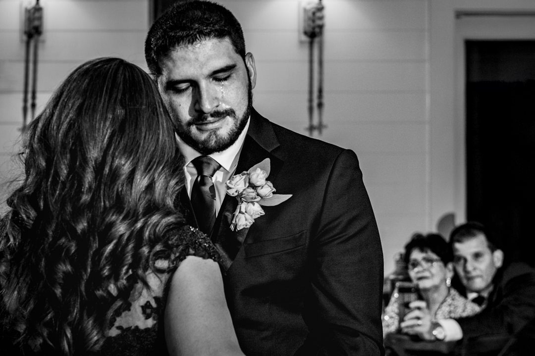 Groom with tears streaming face while dancing with mother during their dance. Father and grandmother watching in background.