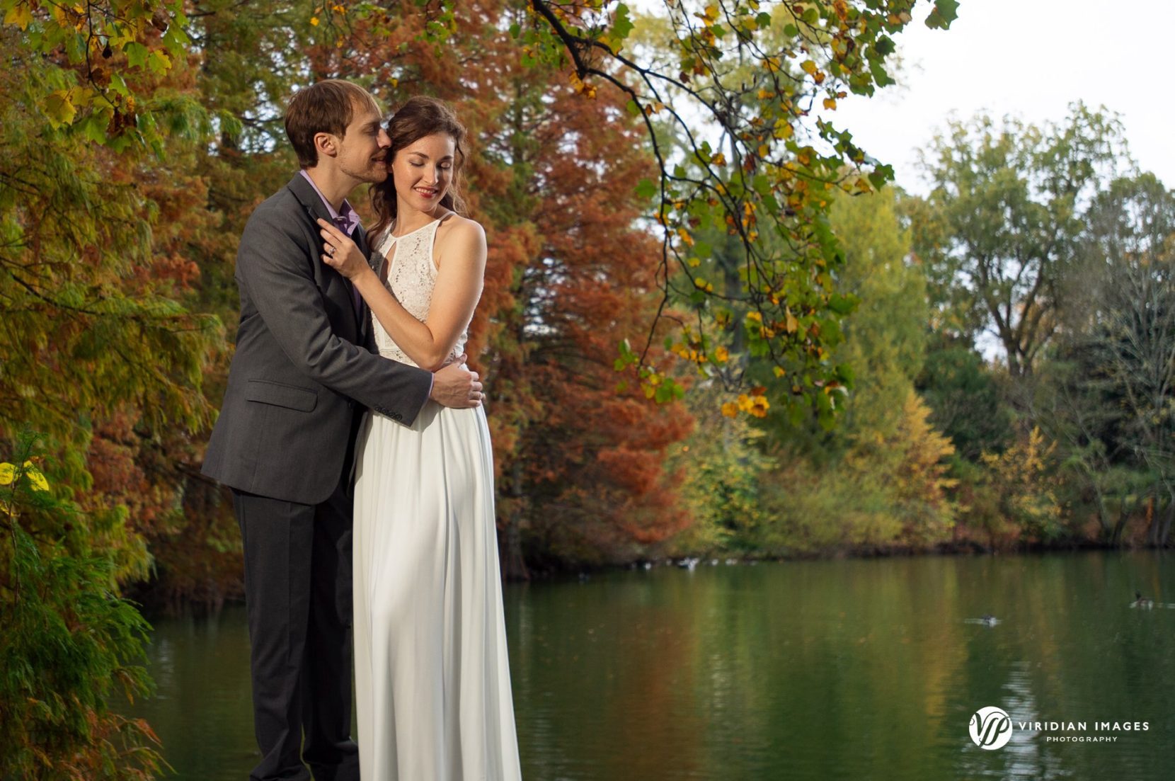 couple embrace along tree lined fall foliage and lakeside at Piedmont Park